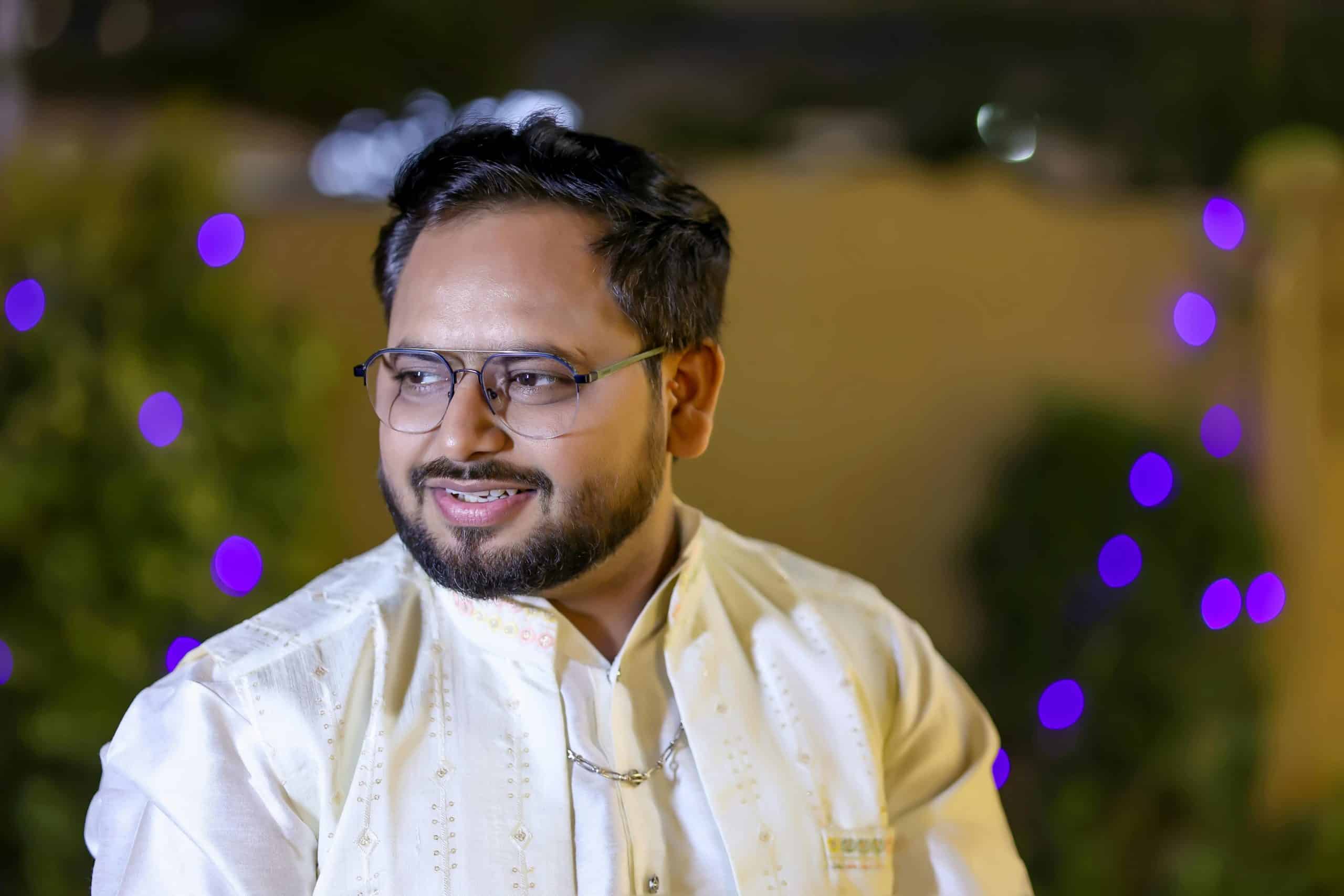 A man in traditional attire smiles with colorful bokeh lights in the background, creating a festive mood.