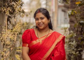 Portrait of a woman wearing a red sari with a soft smile, surrounded by greenery in an outdoor setting.
